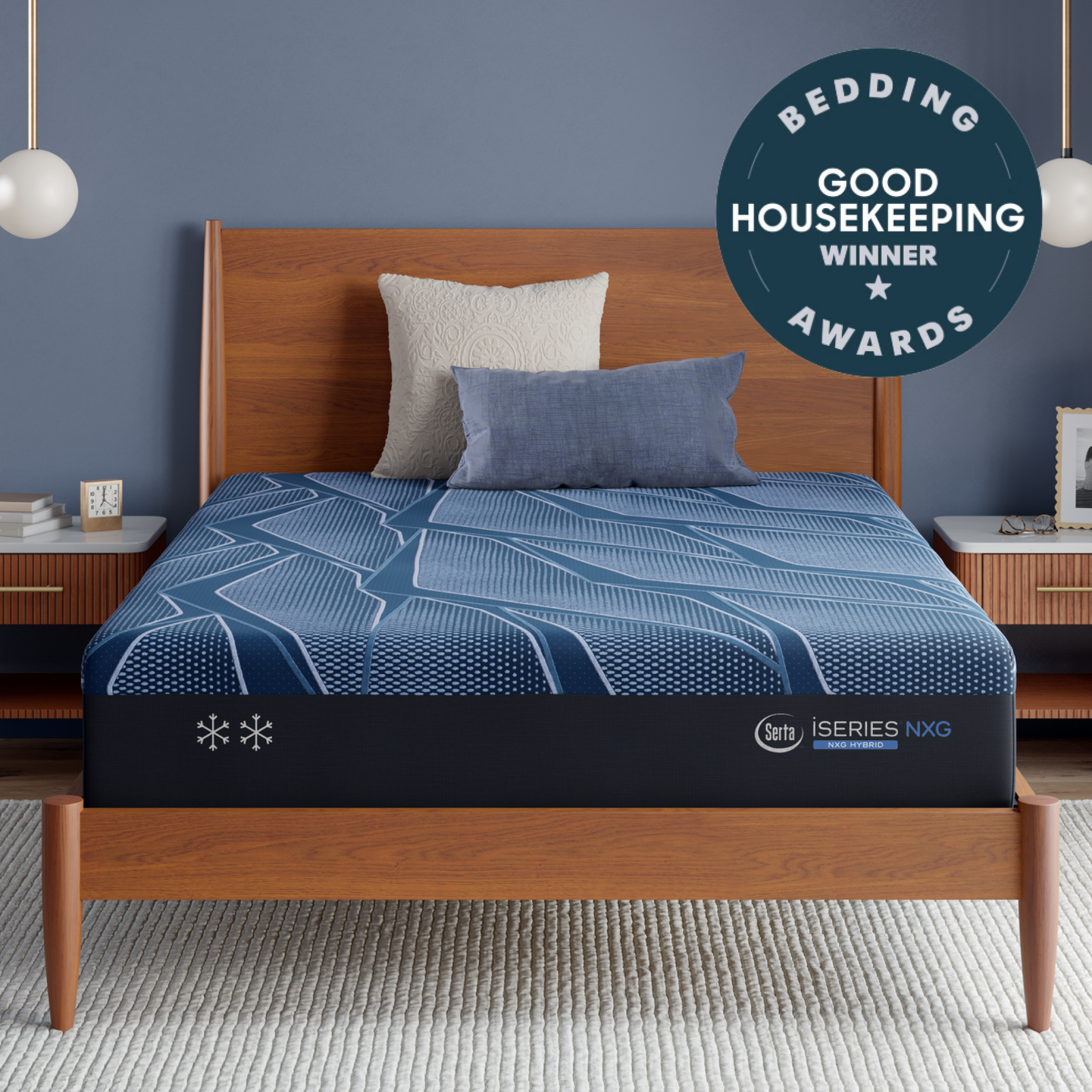 A Blue Serta® iSeries® NXG™ Mattress on a wooden bed frame in a styled bedroom setting, with a blue patterned bedding cover, two pillows, side tables, pendant lights, and a circular badge reading 'Bedding Good Housekeeping Winner Awards.' The mattress brand mark 'iseries' is visible near the bottom edge.