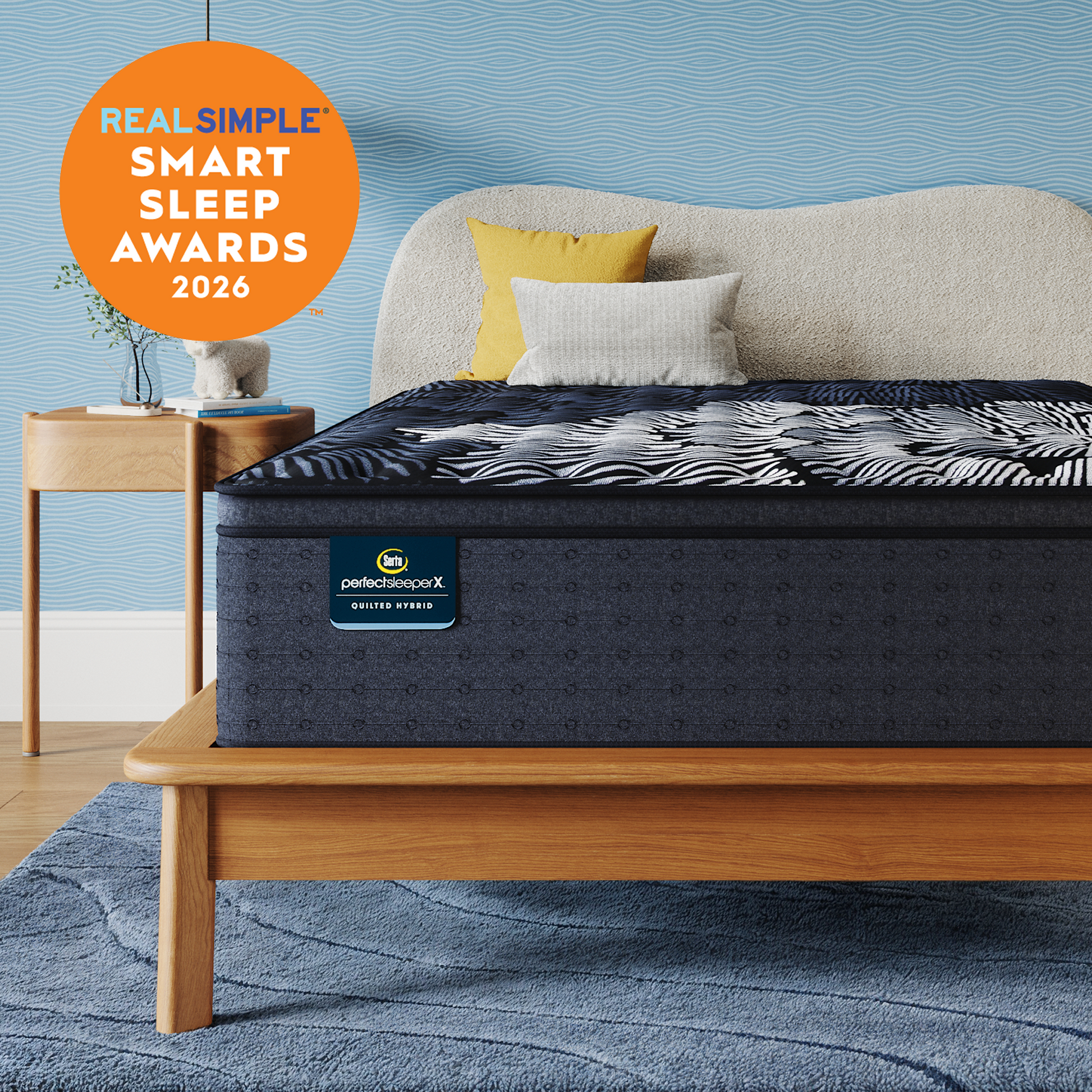 A Queen-sized Perfect Sleeper X Quilted Hybrid mattress with a dark navy and white patterned top surface is shown on a wooden bed frame in a styled bedroom. The background wall is textured light blue, with a beige upholstered headboard, two accent pillows (one yellow, one white), a matching blue rug, and a small wooden side table.